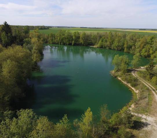 A photo taken from a drone of Juvanzé Blue showing the whole of the lake surrounded by trees.